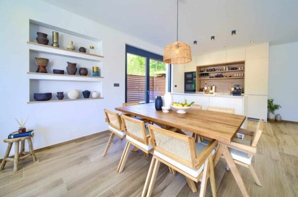 Moderna blagovaonica ima drveni stol okružen laganim stolicama, s ukrasnom keramikom na policama i kuhinjskim aparatima vidljivim u pozadini. Prirodno svjetlo struji kroz velike prozore, dodajući toplinu prostoru.