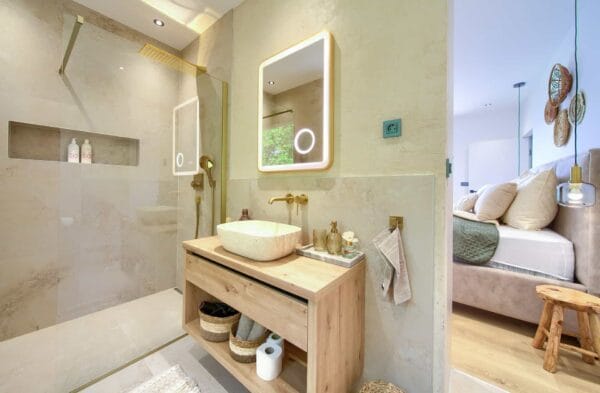 Moderna kupaonica ima stakleni tuš, drveni umivaonik s kamenim umivaonikom i elegantne zlatne elemente. Ugodna spavaća soba vidljiva je kroz otvorena vrata, s istaknutom mekom posteljinom i ukrasnim detaljima.