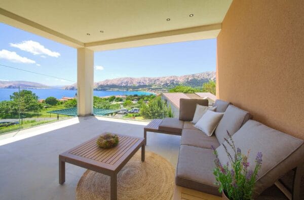 Moderna vanjska terasa s udobnom sofom i drvenim stolom, s pogledom na slikovit pogled na more i planine. U prvom planu vidljivo je bujno zelenilo i kuće.