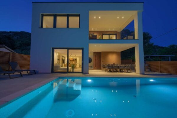 Moderna vila s velikim prozorima, balkonom i terasom smještena u blizini bazena u sumrak. Osvijetljeni interijer ističe ugodan dnevni boravak i blagovaonicu.
